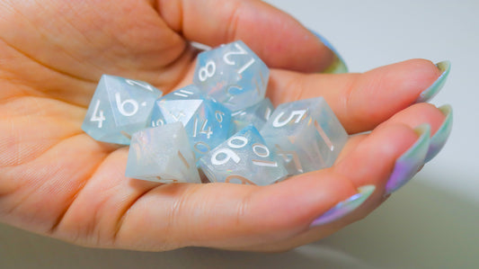 A hand with iridescent sky-blue nails holds a handful of polyhedral dice that match the color.